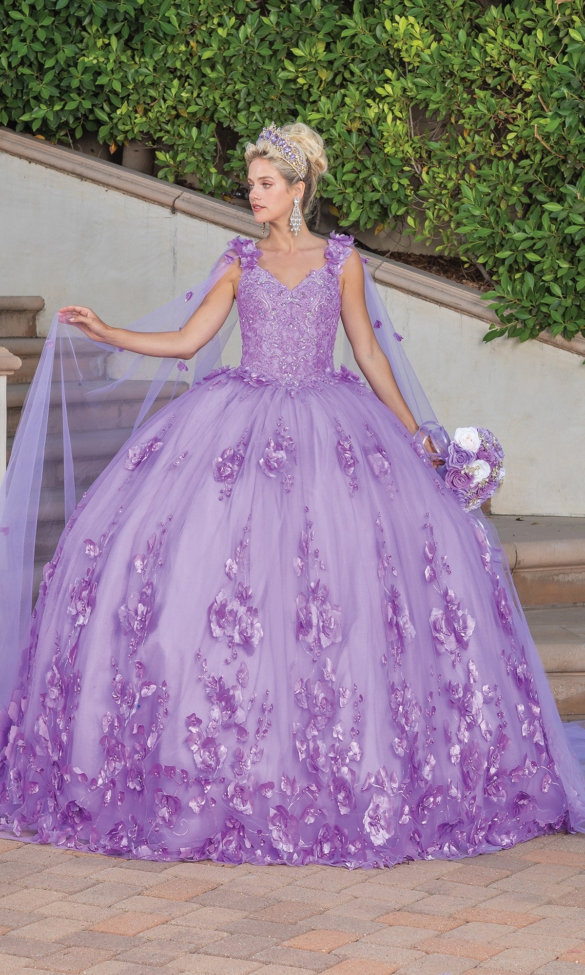 A woman wears the Quinceanera Dress 1695 By Dancing Queen, featuring floral details and an a-line silhouette. She stands on a stone path with a bouquet and tiara, surrounded by lush greenery.